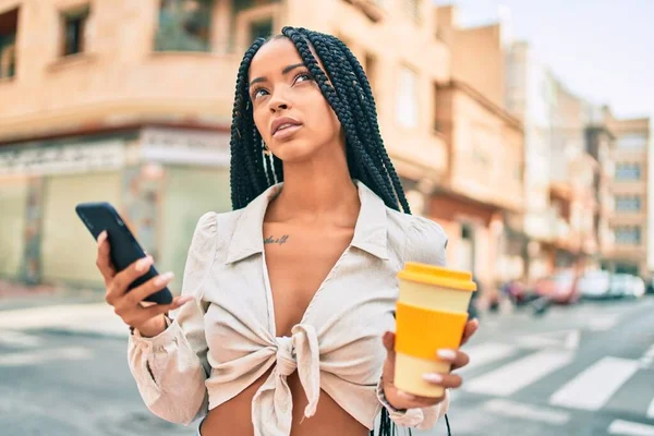 Young african american woman with serious expression using smartphone and drinking coffee at the city.