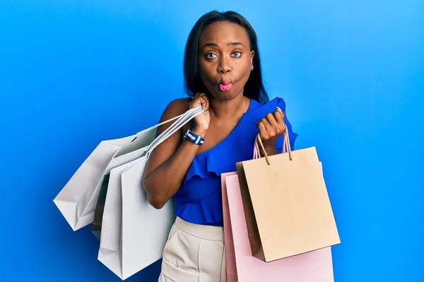 Young african woman holding shopping bags making fish face with mouth and squinting eyes, crazy and comical.
