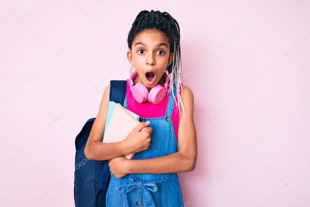 Young african american girl child with braids holding student backpack and books scared and amazed with open mouth for surprise, disbelief face 