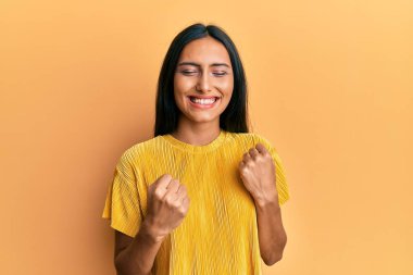 Sarı arka plan üzerine sarı t-shirt giyen genç esmer kadın kolları havada ve gözleri kapalı bir şekilde başarıyı kutluyor ve şaşırıyor. 