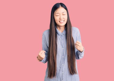 Genç, güzel Çinli bir kadın, gündelik tişört giyip çok mutlu ve heyecanlı bir şekilde kolları havada, gülümseyerek ve başarı için çığlık atarak kazanan bir jest yapıyor. kutlama konsepti. 