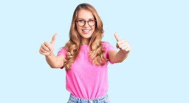 Sarışın, güzel, beyaz bir kadın. Günlük kıyafetler giyiyor ve gözlük takıyor. Elleriyle olumlu jest yapıyor. kazanan jest. 