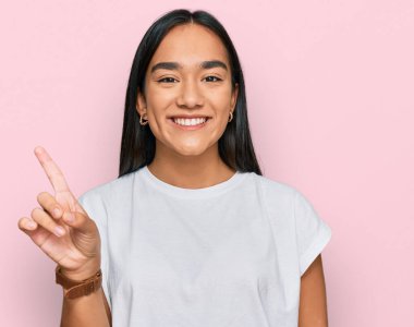 Genç Asyalı kadın, günlük beyaz tişört giyip bir numaralı parmağını gösterirken mutlu ve kendinden emin bir şekilde gülümsüyor.. 