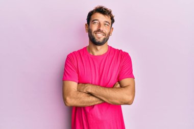 Pembe arka planda pembe tişört giyen sakallı yakışıklı adam kameraya bakarak kollarını kavuşturmuş gülümsüyor. pozitif kişi. 