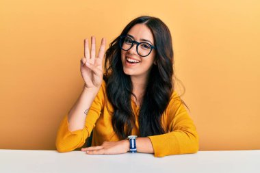 Güzel esmer ve gözlüklü genç bir kadın masanın üzerinde oturuyor ve üç numaralı parmaklarıyla işaret ediyor kendinden emin ve mutlu gülümsüyor.. 