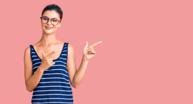 Genç ve güzel bir kadın günlük kıyafetler ve gözlük takıyor gülümsüyor ve kameraya bakıyor iki eliyle ve parmaklarıyla kenarı işaret ediyor. 