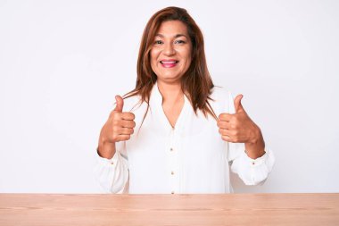 Orta yaşlı esmer, İspanyol iş kadını, gündelik beyaz gömlekli, masa başı tabelasında oturan, elle olumlu jest yapan, gülümseyen ve mutlu bir kadın. Neşeli ifade ve kazanan jest. 