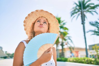 Sarışın, güzel, beyaz bir kadın sıcak bir yaz gününde el fanı kullanarak dışarıda mutlu bir şekilde gülümsüyor.