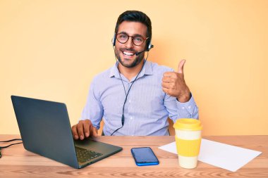 Çağrı merkezindeki operatör kulaklığı takmış, gülümseyen, mutlu ve olumlu gülümseyen, mükemmel ve onay işareti veren İspanyol genç adam. 