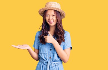 Yazlık şapkalı genç güzel Çinli kız avuç içi gösteriyor ve baş parmaklarıyla iyi bir jest yapıyor, mutlu ve neşeli gülümsüyor. 