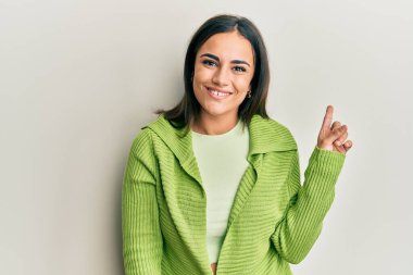 Genç esmer kadın, günlük kıyafetler giyiyor, yüzünde kocaman bir gülümseme var. Elinle kameraya bakıyor.. 