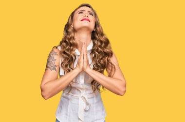 Sıradan kıyafetler giyen genç sarışın kız elleri bağlı yalvarıyor ve dua ediyor. Yüzünde umut ifadesi var. Duygusal ve endişeli. Yalvarıyor. 