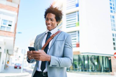 Genç Afrikalı Amerikalı iş adamı mutlu bir şekilde gülümsüyor. Yüzünde gülümsemeyle şehrin caddesinde akıllı telefon kullanarak dikiliyorsun..