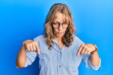 Orta yaşlı, sıradan kıyafetler giyen ve gözlüklü bir kadın üzgün ve üzgün görünüyor, parmaklarıyla yön gösteriyor, mutsuz ve depresif.. 