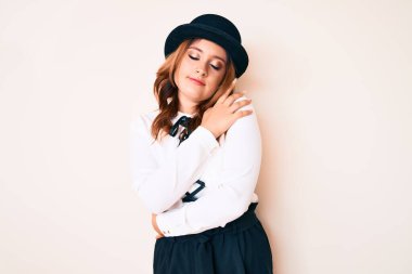 Zarif bir tarzı ve şapkası olan güzel beyaz bir kadın mutlu ve olumlu bir şekilde kendine sarılıyor, kendinden emin gülümsüyor. Sevgi ve özgüven. 