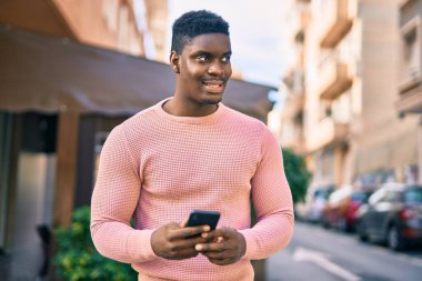 Genç Afrikalı Amerikalı adam şehirde akıllı telefon kullanarak mutlu bir şekilde gülümsüyor..