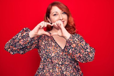 Genç, güzel, kızıl saçlı, çiçek desenli bir elbise giyen, kırmızı arka planda duran, aşkla gülümseyen, elleriyle kalp sembolü yapan bir kadın. Romantik konsept.