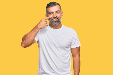 Orta yaşlı, beyaz tişört giyen yakışıklı bir adam, el parmağı ve burnu olan neşeli bir şekilde gülümseyen bir adam. güzellik konsepti 