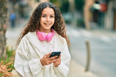 Güzel İspanyol kız şehirde akıllı telefon ve kulaklık kullanarak mutlu bir şekilde gülümsüyor..