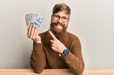 Genç İrlandalı kızıl saçlı adam masanın üzerinde oturmuş gülümsüyor mutlu bir şekilde eli ve parmağı ile işaret ediyor. 