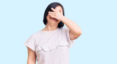 Down sendromlu esmer kadın, sıradan beyaz tişörtler giyiyor, gülümsüyor ve elleri yüzünde şaşkınlıktan gözleri kapatıyordu. kör kavram. 