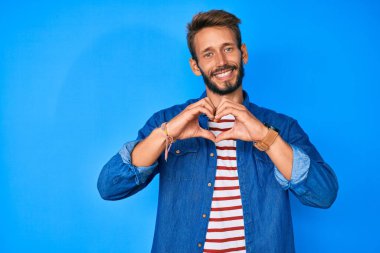 Sarışın, sakallı, sıradan kıyafetler giyen, aşık bir şekilde gülümseyen elleri ile kalp sembolü yapan beyaz bir adam. romantik kavram. 