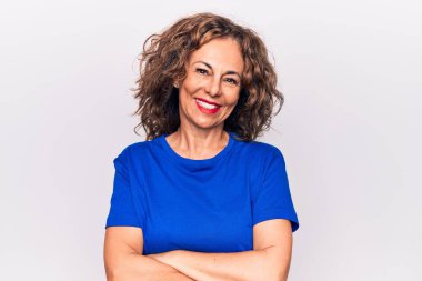 Orta yaşlı, esmer, mavi tişörtlü güzel bir kadın beyaz arka planda durup kollarını kavuşturup kameraya bakarken gülümsüyor. Pozitif kişi.