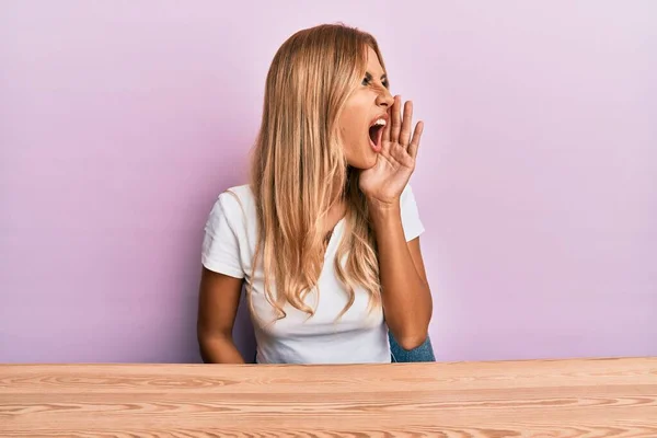 美しいブロンドの若い女性がカジュアルな服を着てテーブルの上に座って大声で叫び 口の中で手で横に大声で叫ぶ コミュニケーションの概念 — ストック写真