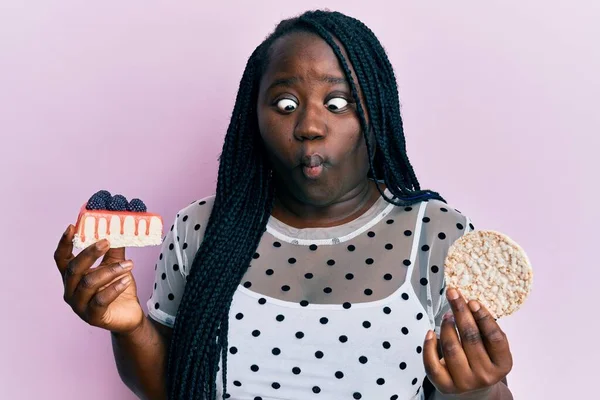 Young black woman with braids eating cake slice and rice crackers making fish face with mouth and squinting eyes, crazy and comical.