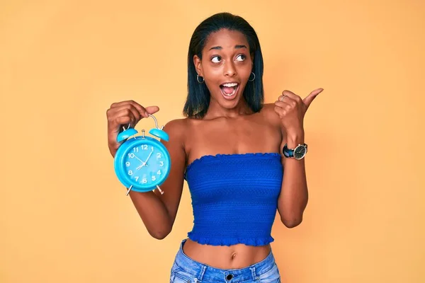 Joven Afroamericana Chica Sosteniendo Despertador Apuntando Pulgar Hacia Lado Sonriendo —  Fotos de Stock