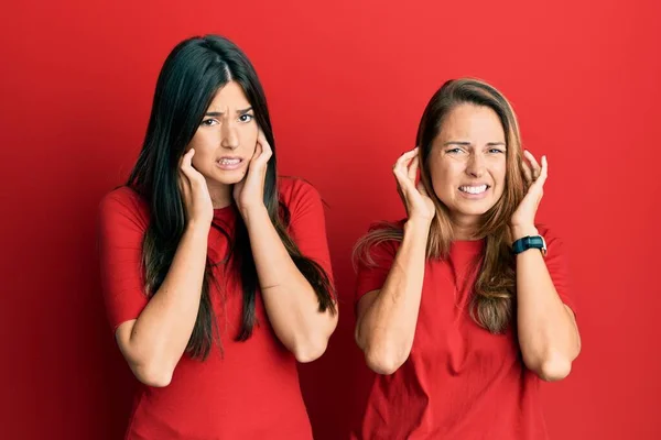 赤い背景にカジュアルな服を着た母親と娘のヒスパニック系の家族は 大きな音楽のノイズのためのいらいらした表情で指で耳をカバーしています 聴覚障害の概念 — ストック写真