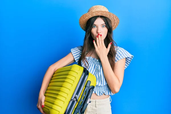 Young beautiful caucasian girl wearing summer dress and holding cabin bag covering mouth with hand, shocked and afraid for mistake. surprised expression