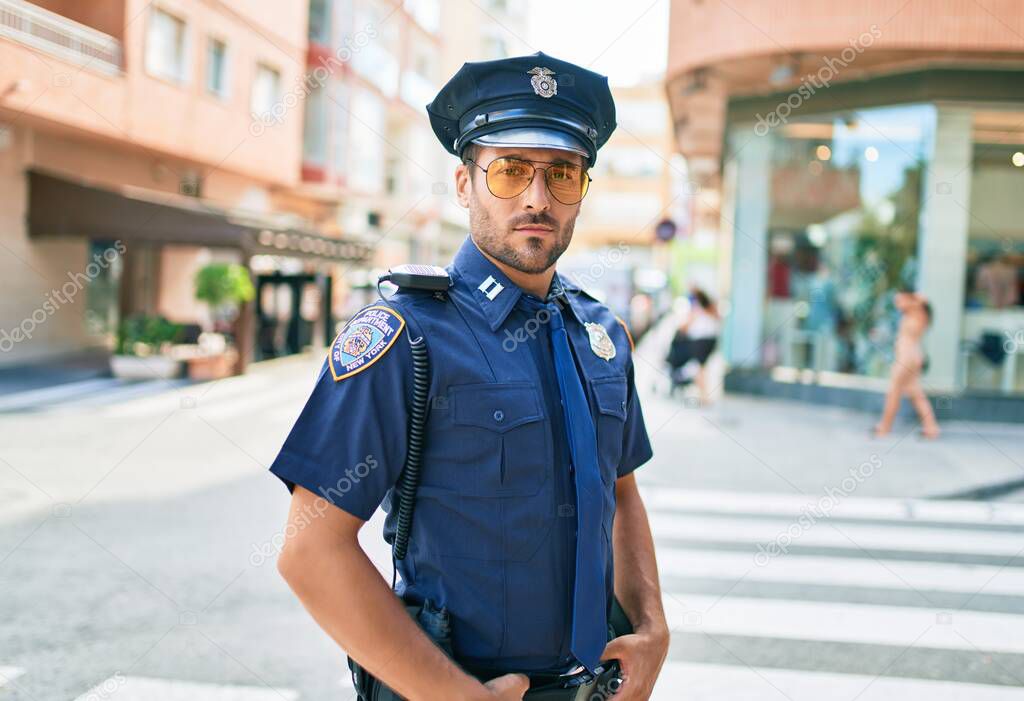 Young handsome hispanic policeman wearing police uniform. Standing with serious expression at town street.
