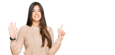 Genç esmer kadın günlük kışlık süveter giyer ve yedi numaralı parmaklarıyla işaret ederken kendinden emin ve mutlu bir şekilde gülümser.. 