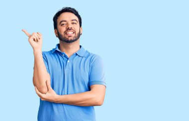 Yüzünde kocaman bir gülümseme olan, gündelik kıyafetler giyen genç İspanyol adam, kameraya el ve parmak ile bakıyor.. 