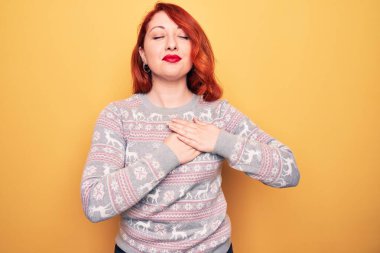 Güzel kızıl saçlı kadın, sarı arka planda ren geyiği olan bir süveter giyiyor. Elleri göğsünde gülümsüyor, yüzünde minnet dolu bir jest ile gözleri kapalı. Sağlık kavramı.