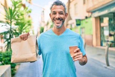 Orta yaşlı, kır saçlı bir adam elinde kahveyi alıp şehir caddesinde içinde yiyecek olan kağıt torbaları dağıtıyor..