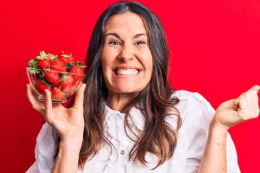 Genç, güzel esmer kadın kırmızı arka planda bir kase sağlıklı çilek tutuyor. Gururla bağırıyor, zaferi kutluyor ve kollarını kaldırarak başarıyı kutluyor.