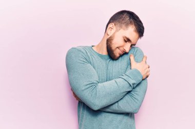 Genç yakışıklı bir adam sıradan bir süveter giyip kendine sarılıyor mutlu ve pozitif, kendinden emin gülümsüyor. Sevgi ve özgüven. 