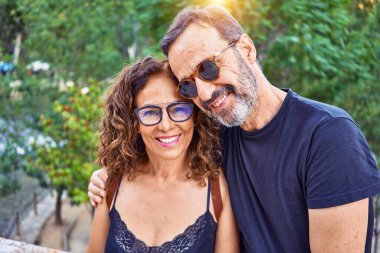 Orta yaşlı bir çift, taş duvarda oturup parktaki kameraya bakarken mutlu bir şekilde gülümsüyor.