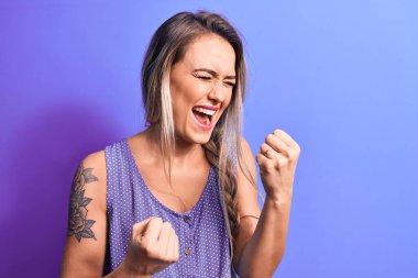 Genç, güzel, sarışın bir kadın. Sıradan bir tişört giyiyor. Mor arka planda dikiliyor.