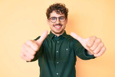 Kıvırcık saçlı, günlük kıyafetler giyen ve gözlük takan genç beyaz adam elinle olumlu jest yapmayı onaylıyor, gülümsüyor ve başarı için mutlu. kazanan jest. 