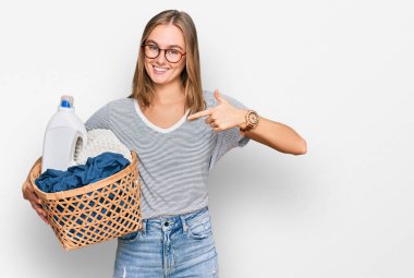 Güzel sarışın kadın çamaşır sepeti ve deterjan şişesiyle mutlu ve gururlu bir gülümsemeyi işaret ediyor. 