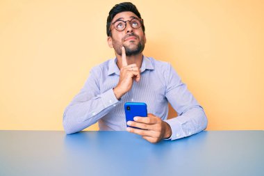 Genç İspanyol bir adam akıllı telefon kullanıyor. Masada oturuyor. Ciddi bir yüz ifadesi var. 