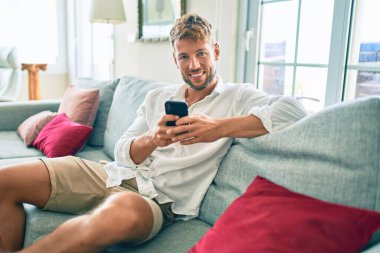 Yakışıklı beyaz adam mutlu gülümsüyor. Evdeki kanepede otururken akıllı telefon kullanıyor.