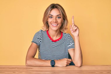 Gündelik giysiler giyen beyaz güzel bir kadın masaya oturmuş başarılı bir fikirle işaret ediyor. Heyecanlı ve mutlu. Bir numara.. 