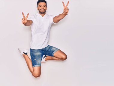 Genç, yakışıklı, sıradan kıyafetler giyen, mutlu gülümseyen bir İspanyol. Yüzünde gülümsemeyle zıplıyor zafer işareti yapıyor parmakları izole beyaz arka planda.