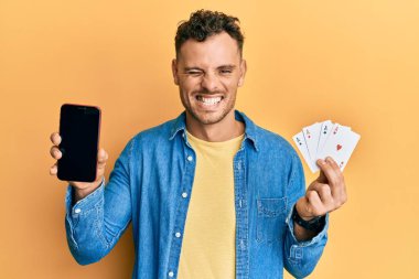 Poker kartları ve akıllı telefon tutan genç İspanyol adam kameraya seksi bir ifadeyle bakıyor, neşeli ve mutlu bir yüz.. 