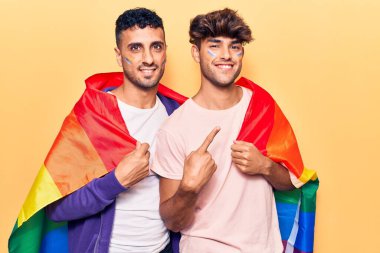 Gökkuşağı lgbtq bayrağı taşıyan genç eşcinsel çift mutlu bir şekilde el ve parmak işareti yapıyor. 