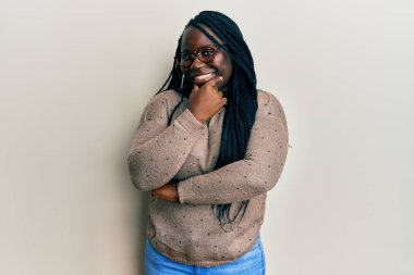 Örgülü, sıradan kıyafetler ve gözlüklü genç siyahi kadın kameraya kendinden emin bir şekilde bakıyor kollarını kavuşturmuş ve çenesini kaldırmış gülümsüyor. Olumlu düşünmek. 